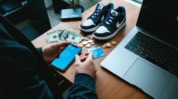 Limited-edition sneakers on a shoebox with cash, coins, and a debit card on a desk, a person’s hands hovering near a smartphone, dramatic side lighting, blurred apartment background.