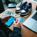 Limited-edition sneakers on a shoebox with cash, coins, and a debit card on a desk, a person’s hands hovering near a smartphone, dramatic side lighting, blurred apartment background.